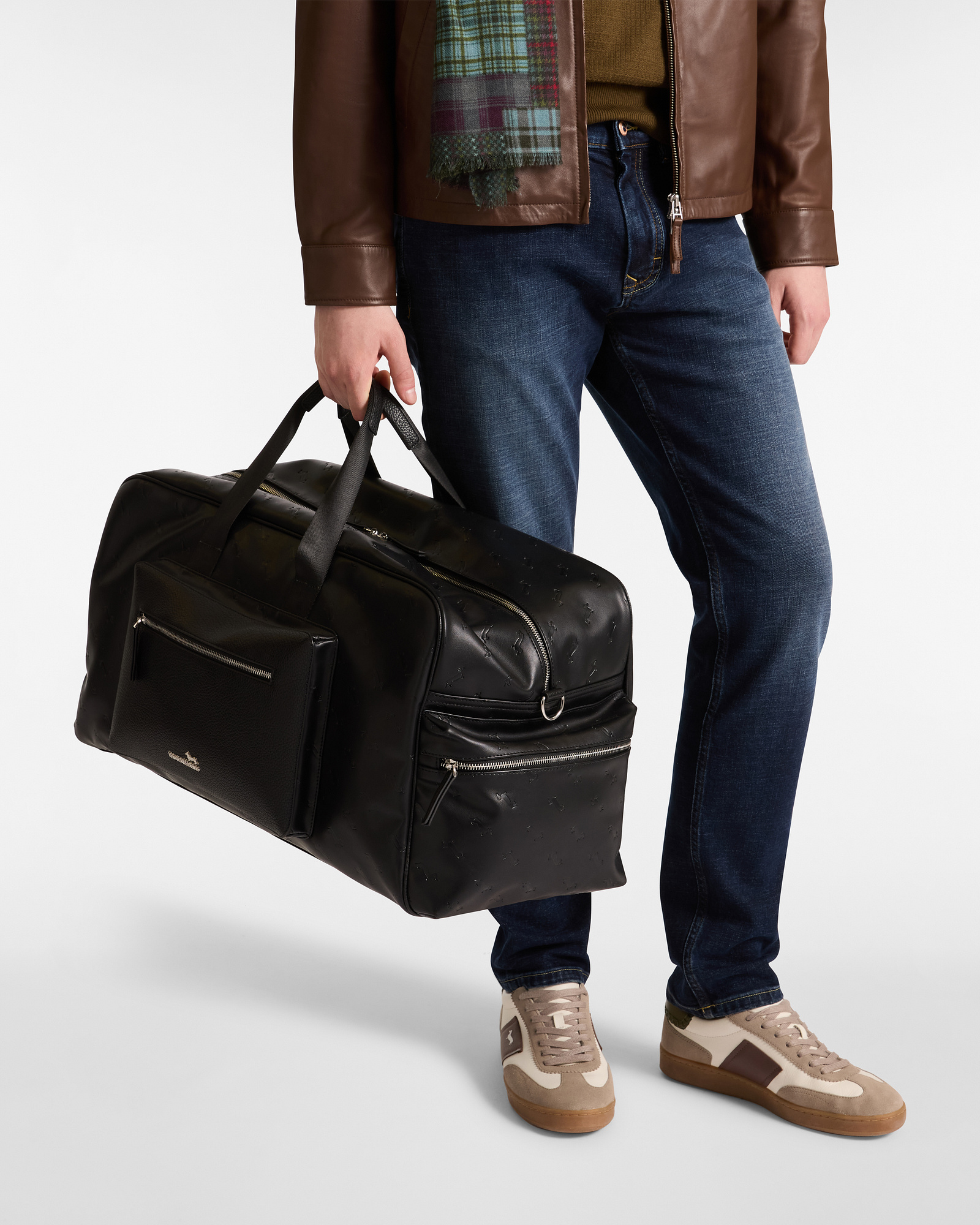 Uomo con borsone nero in pelle, giacca marrone in pelle, jeans e sneakers beige